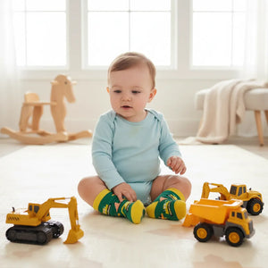Baby sitting on a light-coloured floor with toy construction vehicles and colourful green and yellow digger socks.